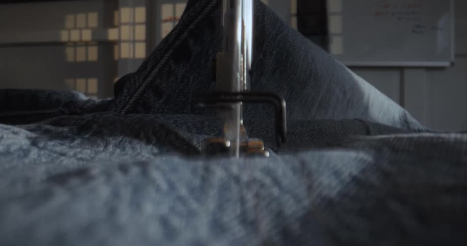 A sewing machine stitches a seam on blue denim fabric in a dramatic, low-angle close-up shot