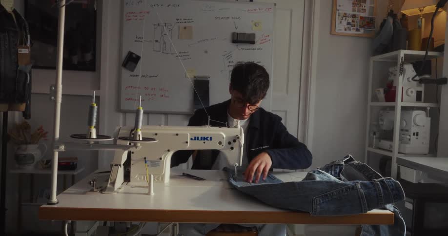 A young male designer carefully sews a pair of denim jeans in his workshop with a Juki sewing machine