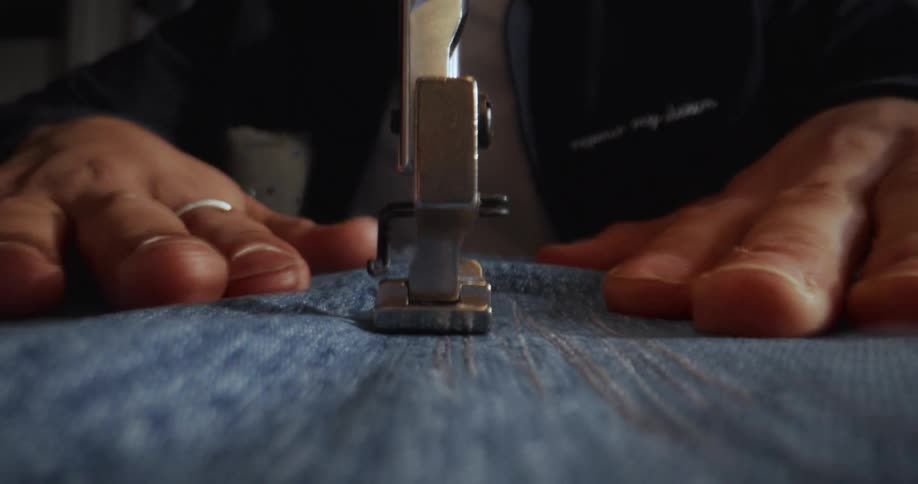 A detailed macro shot captures a person's hands skillfully guiding blue denim fabric through a sewing machine