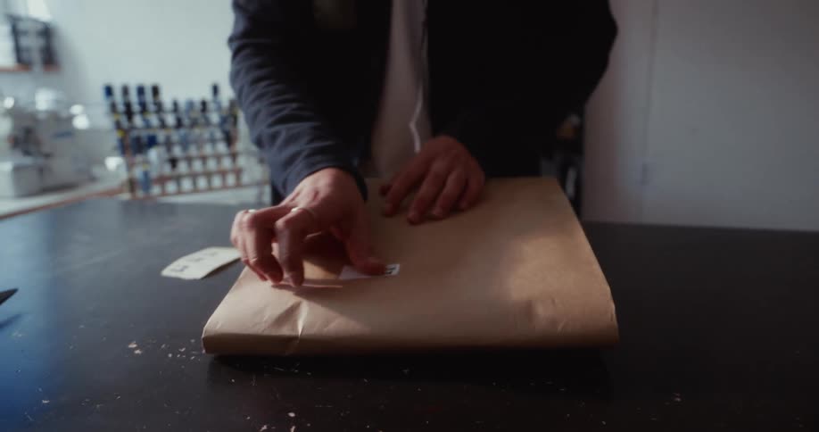 A tailor carefully packages an order in brown paper, adds a sticker, and writes on the label in his workshop
