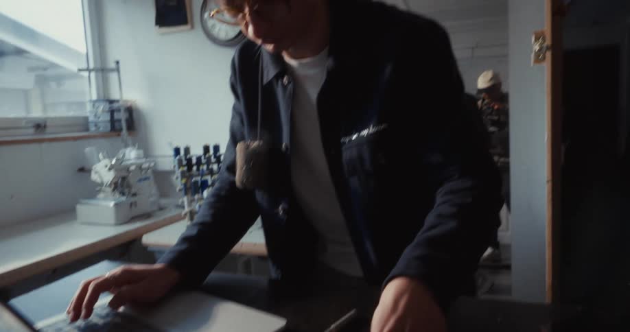 A young fashion designer works on a laptop at his desk in a sunlit creative workshop