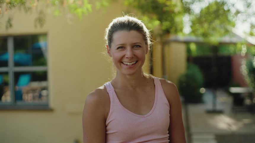 A happy, beautiful woman with her hair in a bun smiles warmly while standing in her sunny backyard