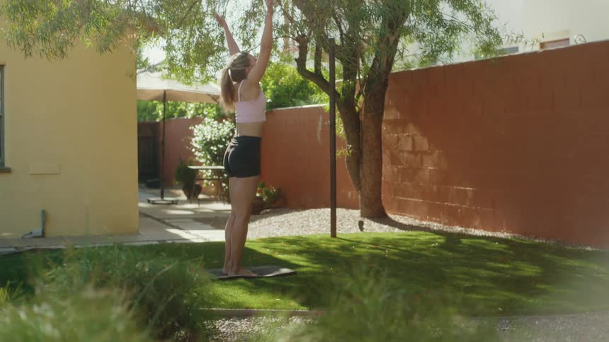 A young woman practices yoga poses outdoors in her sunny backyard on a bright morning