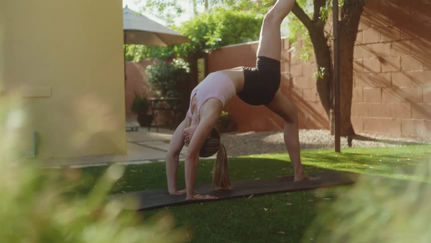 A fit woman gracefully holds an advanced one-legged wheel yoga pose on a mat in her serene, sun-drenched backyard