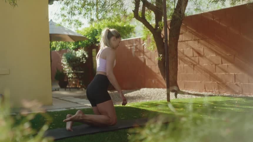 In slow motion, a woman walks barefoot to her yoga mat in a sun-drenched backyard and settles into a calming child's pose