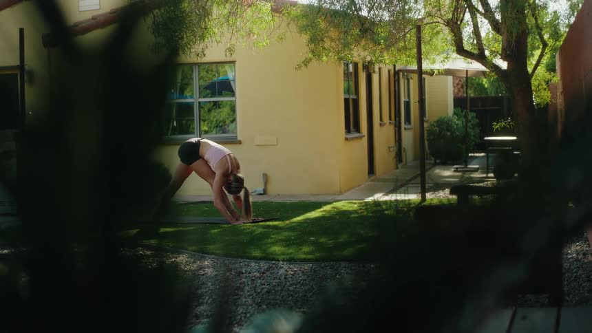 A woman in athletic wear holds a deep yoga stretch in her sunny backyard as seen from behind a bush
