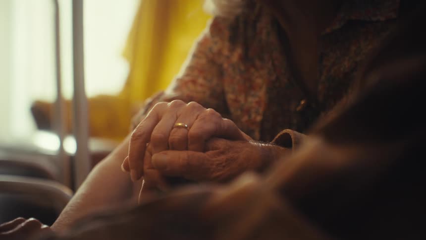 A close up of an elderly couple's hands clasped together as they look at an old photo album, reminiscing