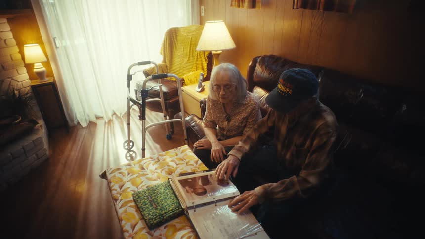 An elderly couple sits together in their dimly lit living room, fondly looking through a family photo album and reminiscing about the past