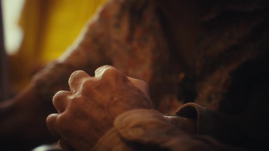 An intimate close-up shot of an elderly couple's weathered hands holding each other in a tender embrace