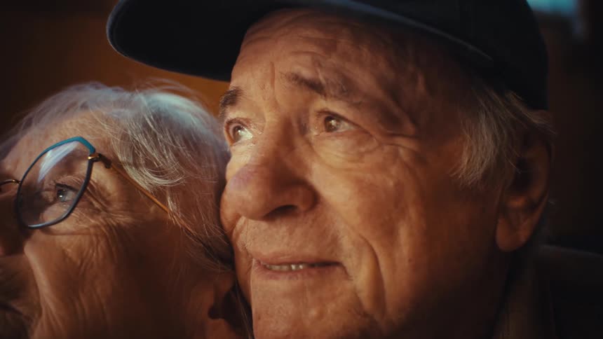 A heartwarming close-up of an elderly couple, with the man wearing a 'World's Best Grandpa' hat, watching something off-screen with expressions of joy and wonder