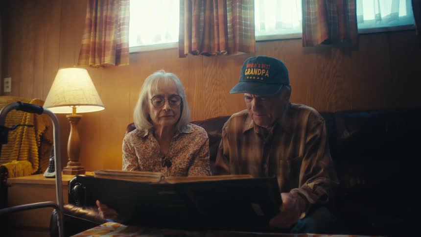 An elderly couple sits on their couch and looks through a photo album, reminiscing about their past as the camera slowly zooms in on the woman's face