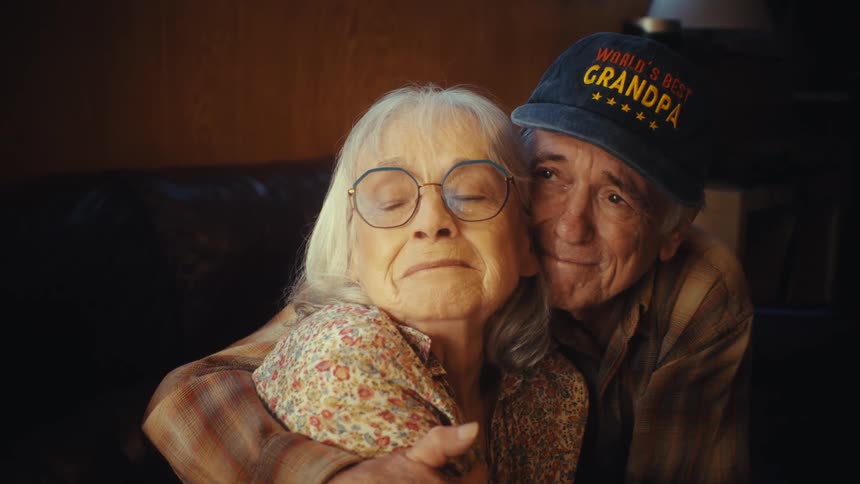 An affectionate elderly man wearing a 'World's Best Grandpa' hat lovingly hugs his wife as they sit together on a couch at home