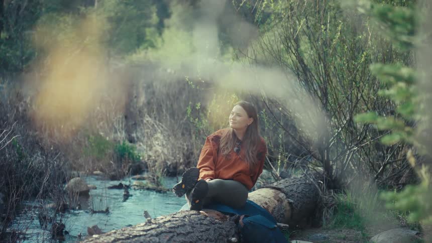 A serene woman in an orange sweatshirt sits peacefully on a fallen log, enjoying the tranquility of a sun-dappled forest stream