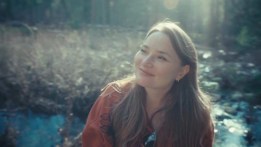 A serene young woman smiles peacefully with her eyes closed, enjoying the warm sunlight and fresh air in a tranquil forest