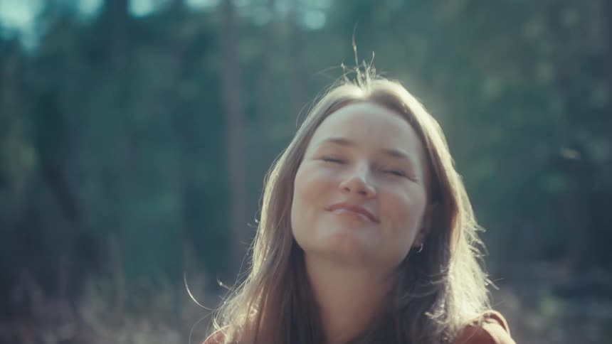 A young woman with a peaceful smile enjoys the warm sunlight shining on her face in a tranquil forest setting