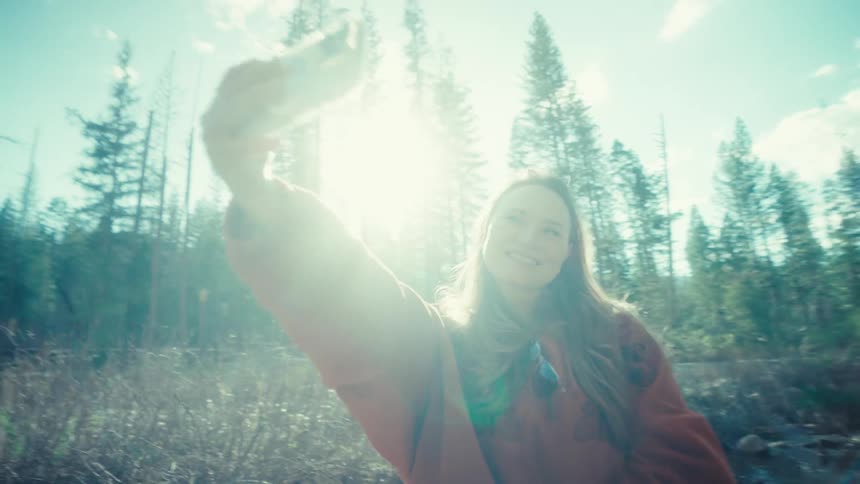 Smiling woman records a video selfie while sitting on a fallen tree in a beautiful sunny forest