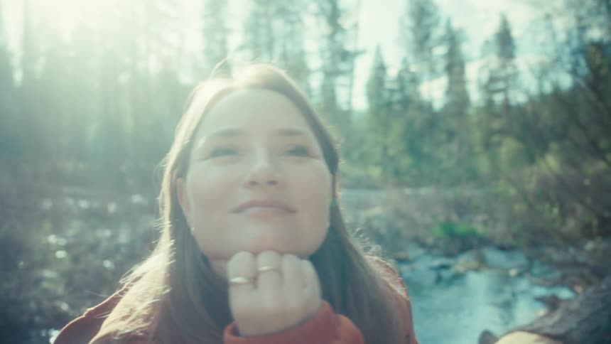 A serene woman with her eyes closed smiles peacefully while basking in the warm sunlight of a tranquil forest