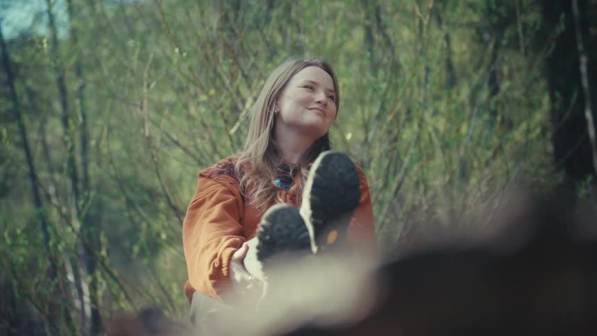 A serene young woman smiles peacefully as she relaxes with her feet up on a log in a sunlit forest