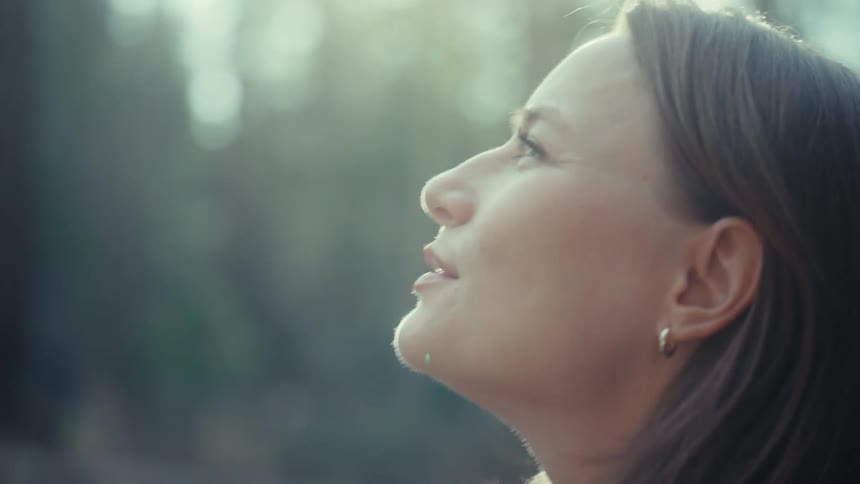 A serene woman smiles peacefully as she looks up towards the golden sunlight in a tranquil forest setting