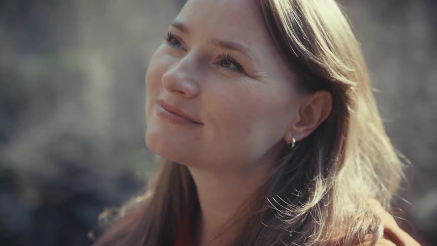 A serene woman with long hair smiles peacefully while enjoying the sunlight in a tranquil outdoor setting