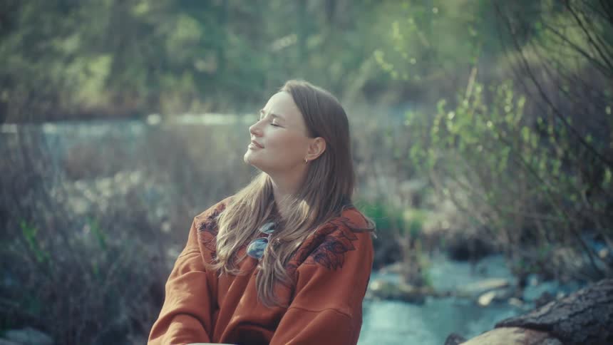 A peaceful woman sits on a log by a forest stream, closing her eyes and smiling as she enjoys the warm sunlight