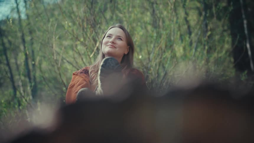A woman wearing hiking boots relaxes with her feet up, smiling peacefully in a sun-dappled forest