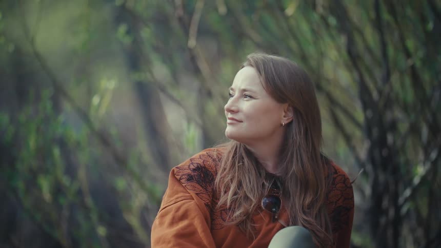 A contemplative woman with long hair gently smiles while sitting and relaxing in a serene natural setting