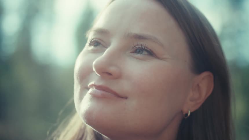 A serene woman with beautiful blue eyes smiles peacefully as she closes them to feel the warm sunlight on her face in a tranquil forest