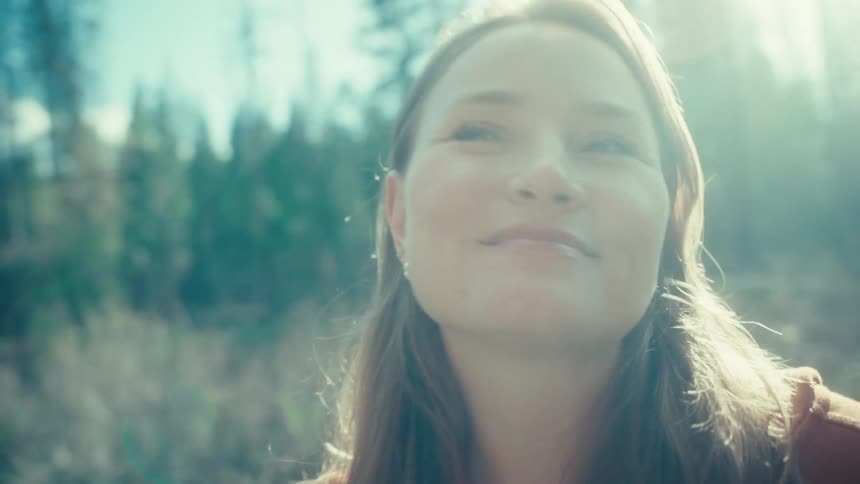 A serene woman smiles as she soaks in the bright, hazy sunlight in a peaceful forest clearing