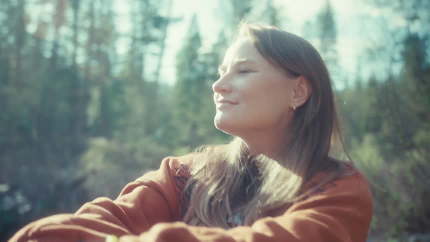 A serene woman sits on a log in a sun-drenched forest, closing her eyes to peacefully soak in the warm sunlight