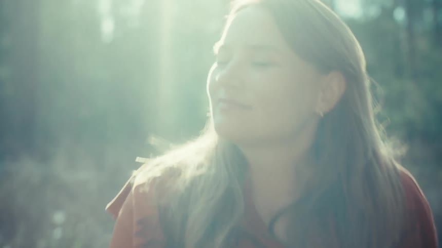 A slow zoom reveals a close-up of a peaceful woman smiling as she enjoys the warm sunlight in the forest