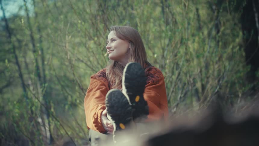 A serene woman in an orange sweater and hiking boots sits and relaxes in a beautiful green forest, smiling as she enjoys nature