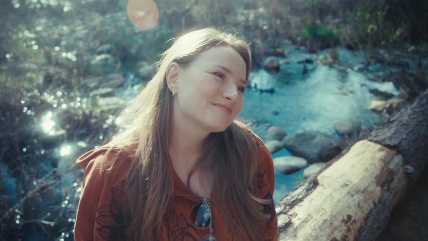 A serene woman sits on a log by a sunlit creek in the woods, smiling as she lifts her face to the warm sunshine