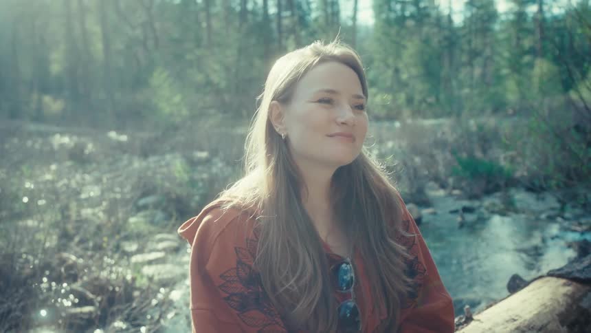 A serene woman finds a moment of peace and tranquility while sitting on a log by a sun-drenched forest stream
