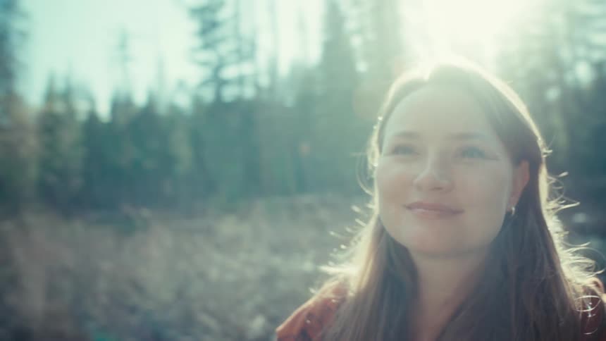 A serene woman with long hair smiles contentedly as she enjoys the warm sunlight in a beautiful forest setting