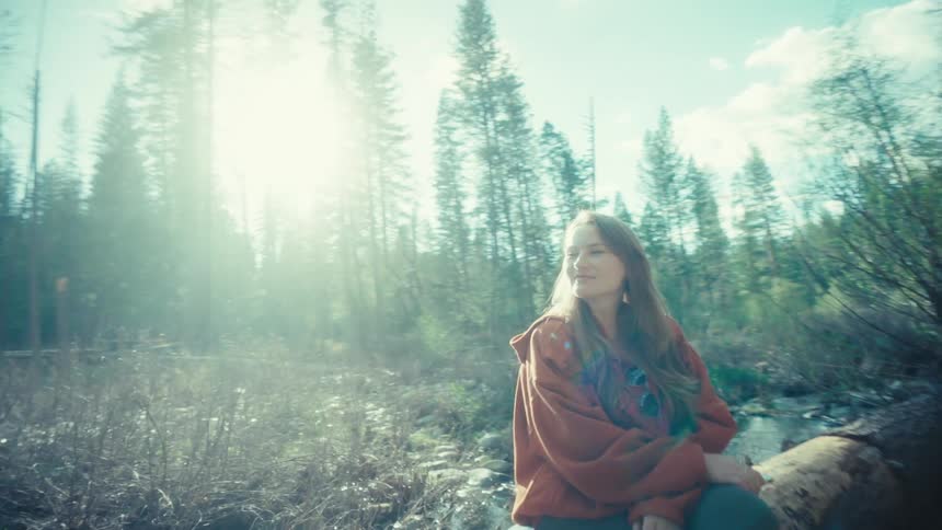 A young woman puts her phone away and soaks in the bright sunshine while sitting on a fallen log in a peaceful forest