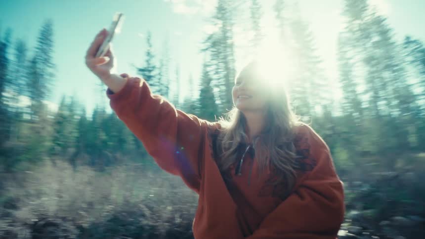 A happy young woman sits on a large fallen log in a sun-flared forest and takes a smiling selfie with her phone