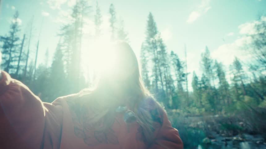 A smiling young woman takes a selfie on her phone while sitting on a log in a sun-drenched forest, enjoying a peaceful moment in nature