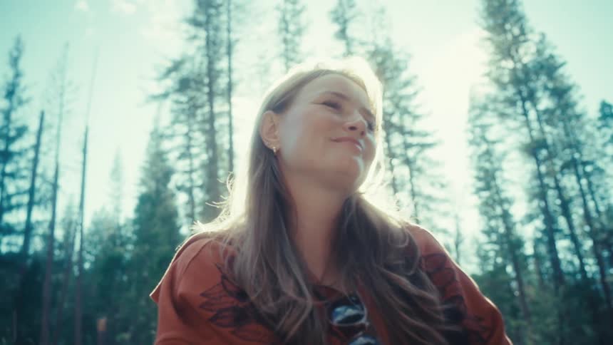 A peaceful young woman sits in a sun-drenched forest, smiling as she enjoys the warm light filtering through the tall trees