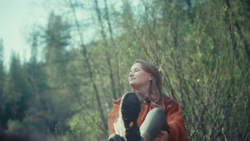 A content young woman in hiking boots takes a peaceful break in a sunlit forest, smiling as she enjoys the serene atmosphere