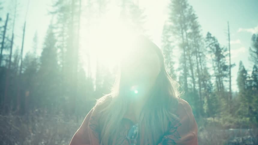 A serene woman enjoys the bright, hazy sunlight in a tranquil forest, looking up with a peaceful smile as the camera tilts towards the sky