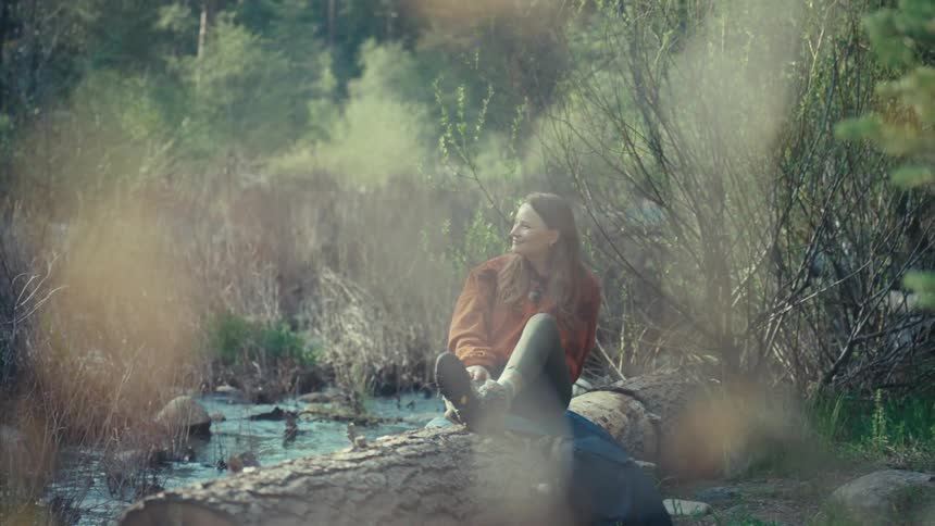 A smiling female hiker rests on a fallen log by a creek, enjoying a peaceful moment in the sun-dappled forest