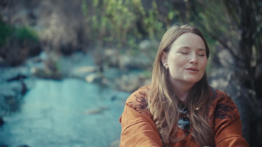 A peaceful woman takes a deep breath and enjoys a tranquil moment while sitting by a flowing stream in the woods