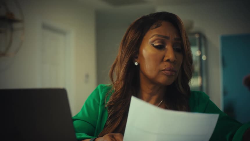 A concerned African American woman sits at her desk and carefully reviews important documents with a serious expression