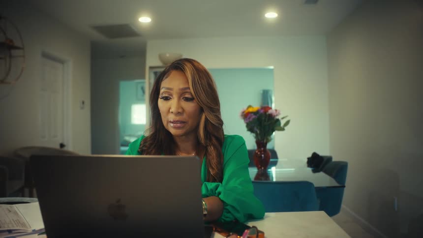 A professional woman in a green blouse works from home on her laptop, her expression shifting from thoughtful to concerned