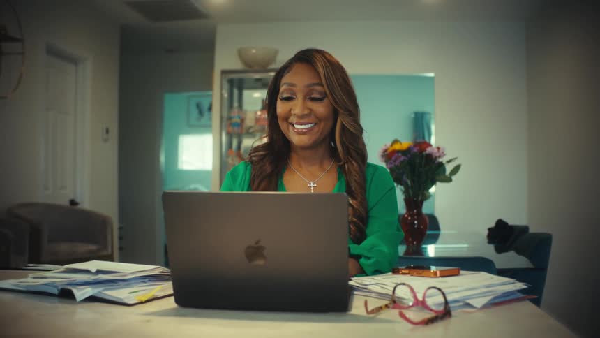 A woman working from home smiles and waves during a video conference call on her laptop in her modern kitchen