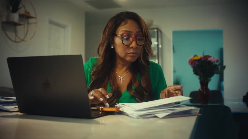 A focused woman in glasses works from home, diligently managing finances on her laptop while reviewing a stack of bills