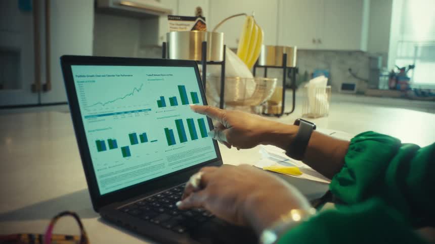 An over-the-shoulder view of a woman working from her kitchen island and pointing at financial performance charts on her laptop