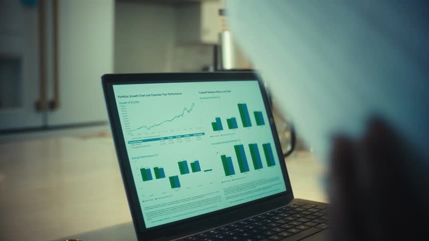 A woman works from home at her kitchen counter, analyzing financial performance charts on her laptop and reviewing paperwork