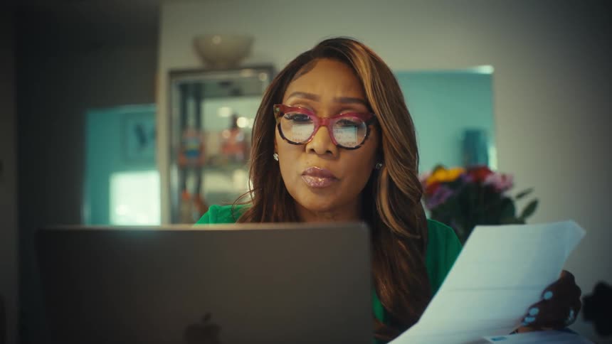 An elegant woman working from home on her laptop reviews a stack of documents, her expression turning serious as she takes off her glasses
